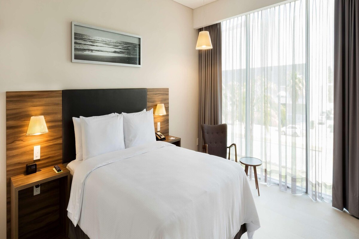 A hotel room with a large bed, two bedside lamps, a nightstand, a chair, a small table, a painting on the wall, and a view through the window.