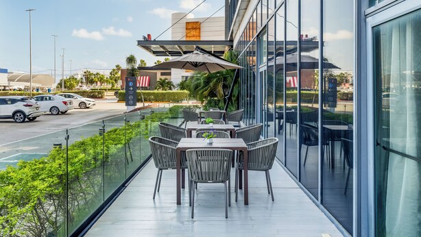 Un área de comedor al aire libre moderna con barandas de vidrio, mesas y sillas, con vista a un estacionamiento y un edificio con una sección naranja característica.