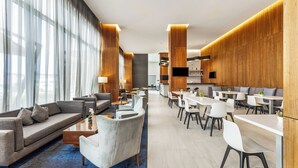 Un moderno vestíbulo de hotel con un sofá, sillones, una mesa de centro, una zona de comedor con mesas y sillas, y una cocina al fondo.