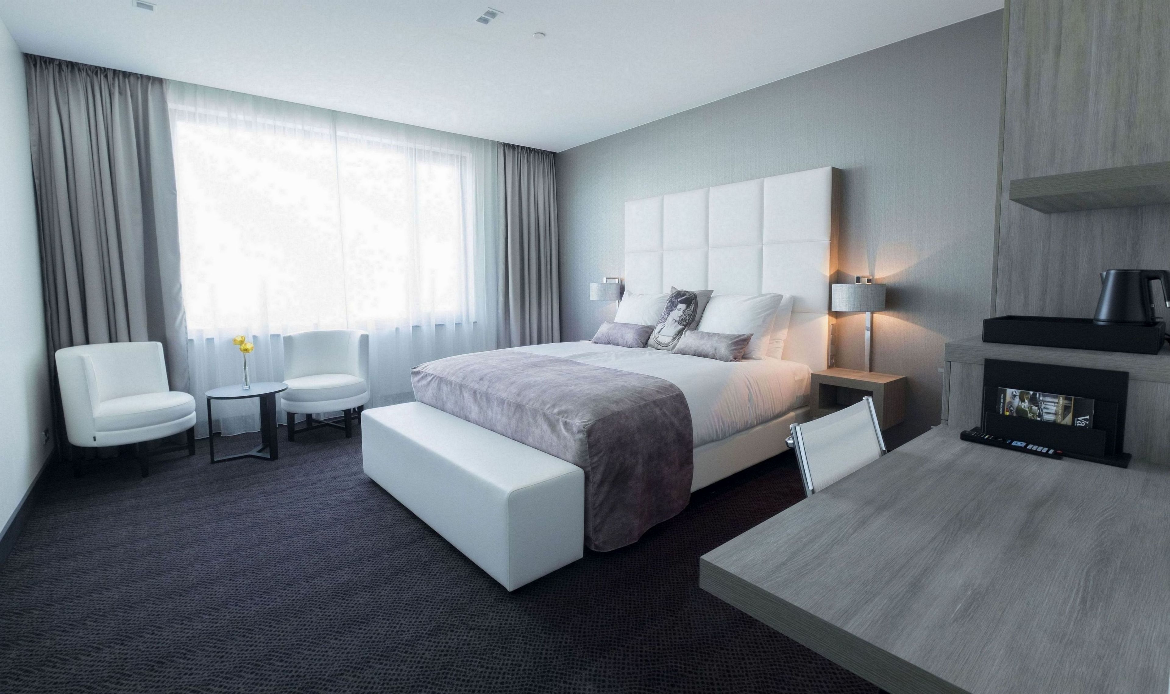 A modern hotel room with a large bed, a bedside table, a chair, a small table, and a TV.