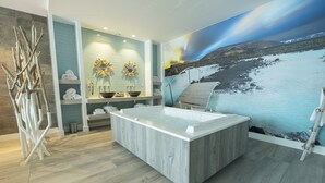 Un baño moderno con una bañera grande, un tocador de madera y un lavabo empotrado en la pared. Hay toallas ordenadas en estantes, y un espejo decorativo con motivos de sol.