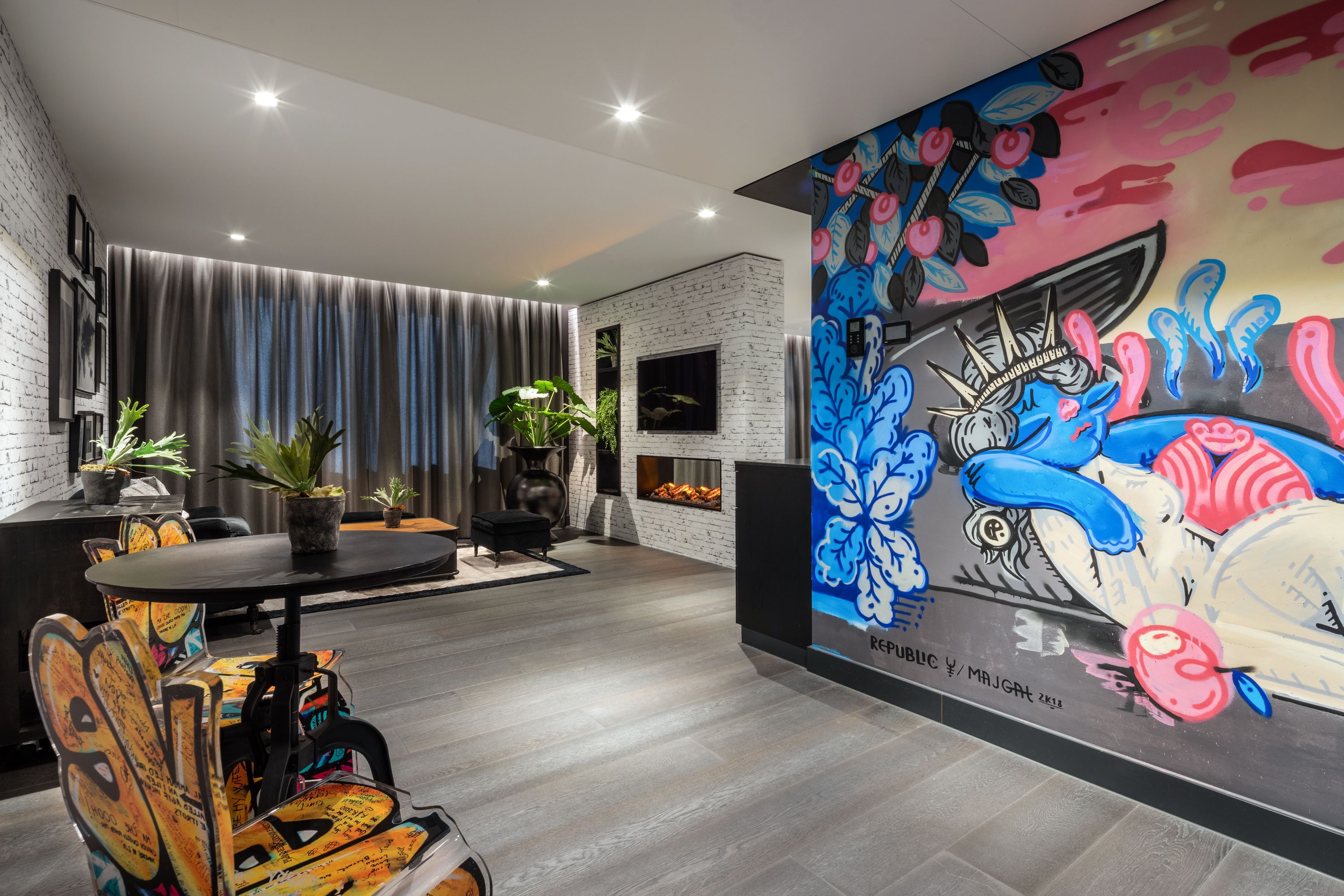 A modern living room with a fireplace, a dining area with graffiti-covered chairs, and a colorful mural on the wall.