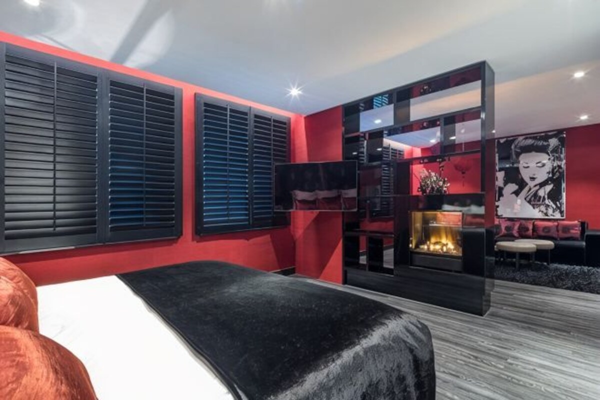 A modern hotel room with a large bed, a fireplace, a red accent wall, and a black and white artwork.