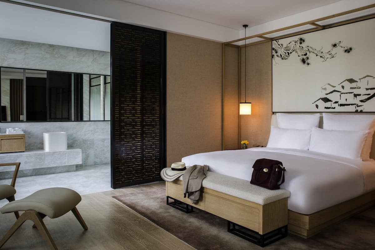A modern hotel room with a large bed, a wooden bench, a desk, and a bathroom with a mirror and sink.