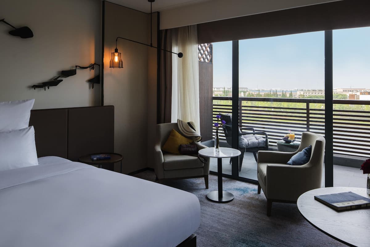 A hotel room with a large bed, a small round table, two armchairs, a sofa, and a balcony with a view of the city.