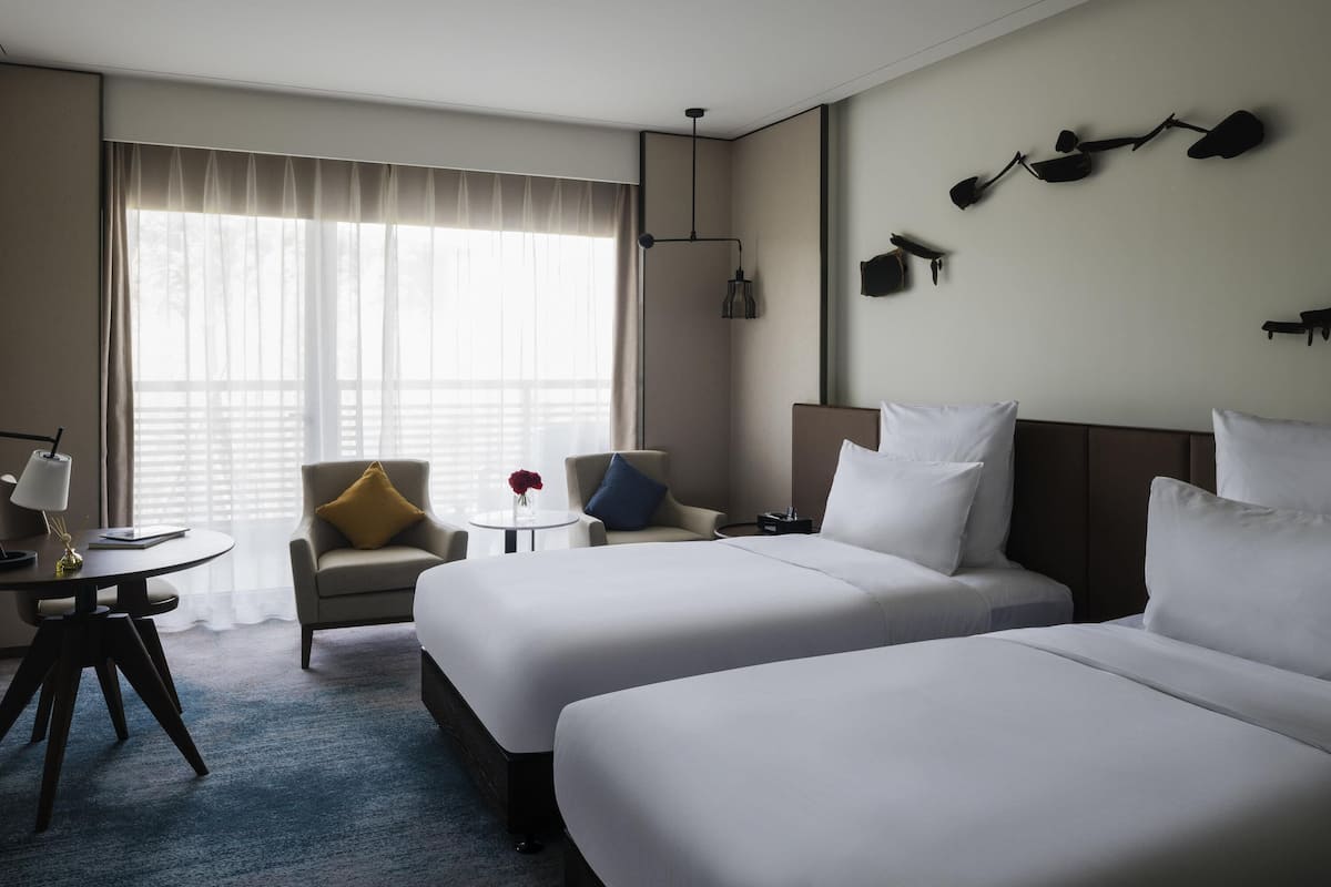 A hotel room with two beds, a desk, a chair, a lamp, and a window with curtains.