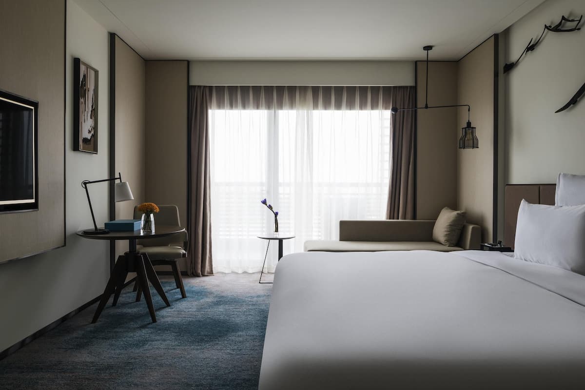 A hotel room with a bed, a desk with a lamp, a chair, a sofa, and a large window with curtains.