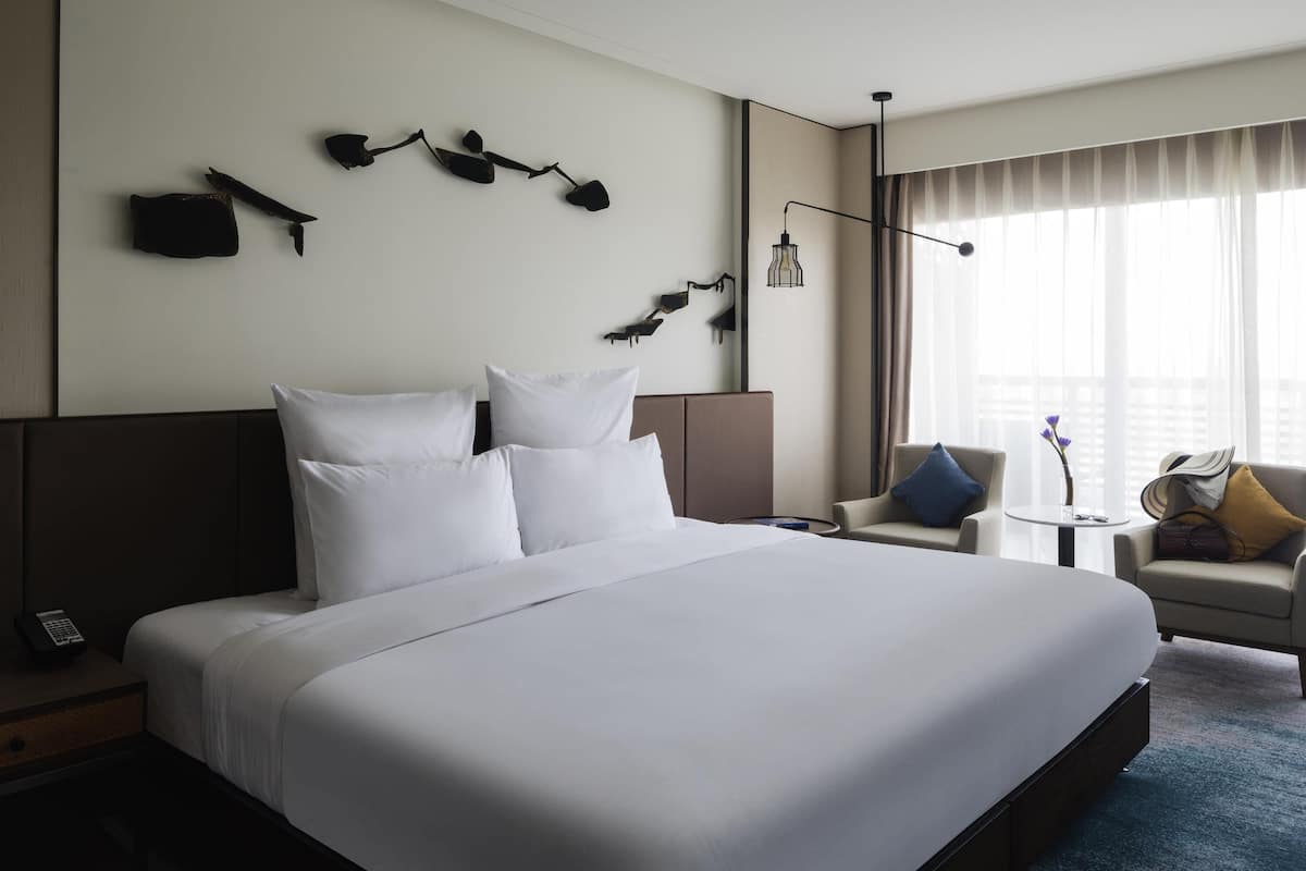 A hotel room with a large bed, a bedside table with a phone, a small round table with a vase, and a sofa with cushions.