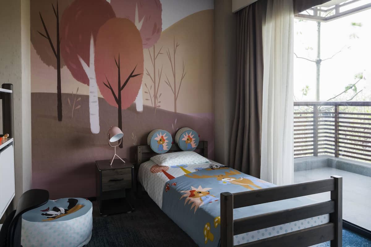 A bedroom with a bed, a nightstand, a lamp, a window with curtains, and a wall mural.