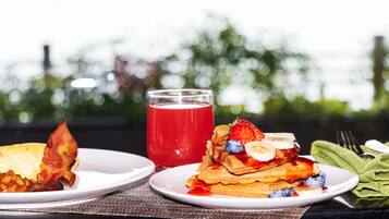 Zwei Teller mit Essen, einer mit Waffeln und Früchten belegt, der andere mit Speck und Bohnen, und ein Glas rotes Getränk auf einem Tisch mit Blick auf Bäume.
