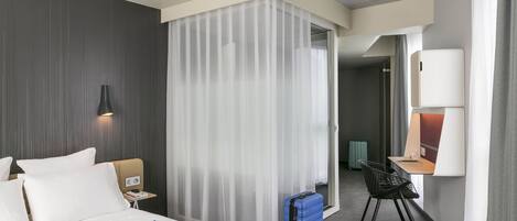 A hotel room with a bed, a desk, a chair, a suitcase, and a lamp.