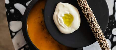 Eine Schüssel Suppe mit einem Klecks Sahne und einem mit Sesamsamen bestreuten Brotstengel auf einem schwarzen Teller, auf einem Terrazoboden platziert.
