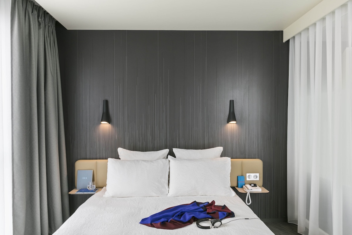 Una habitación de hotel con una cama, dos mesitas de noche, una lámpara y una cortina.