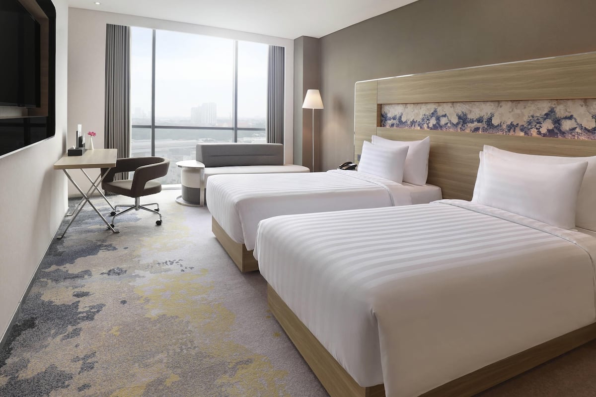 A hotel room with two beds, a desk, a chair, a lamp, and a view of the city.