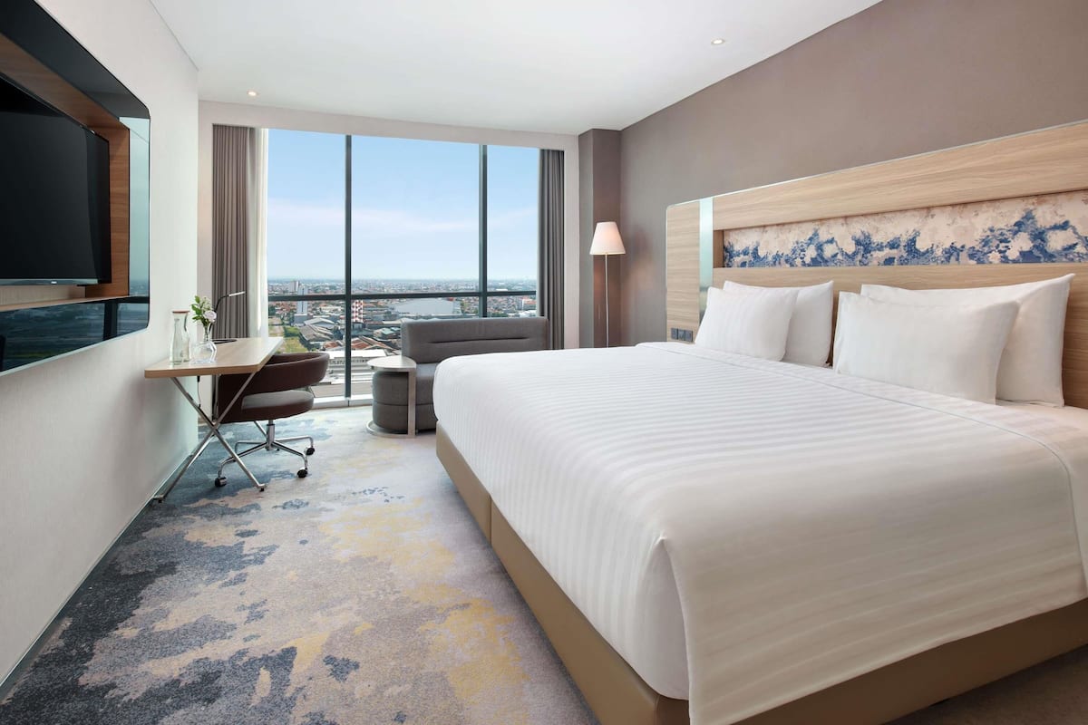 A hotel room with a large bed, a desk with a chair, a TV, and a view of the city.