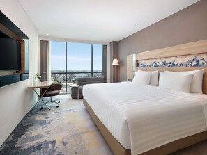 A hotel room with a large bed, a desk with a chair, a TV, and a view of the city.