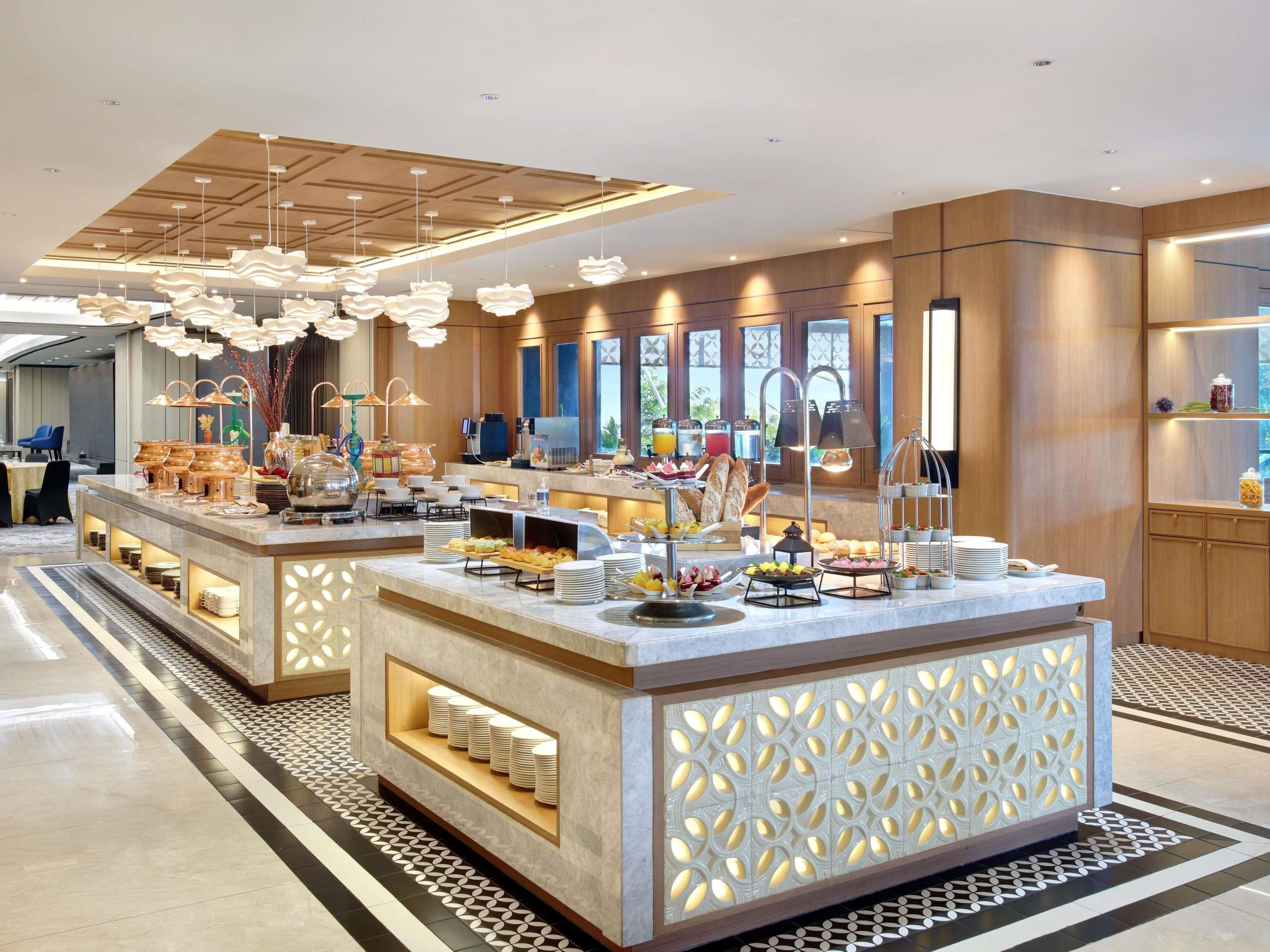 Un área de desayuno de hotel moderna con un bufé central, diversas opciones de asientos y un techo decorativo.