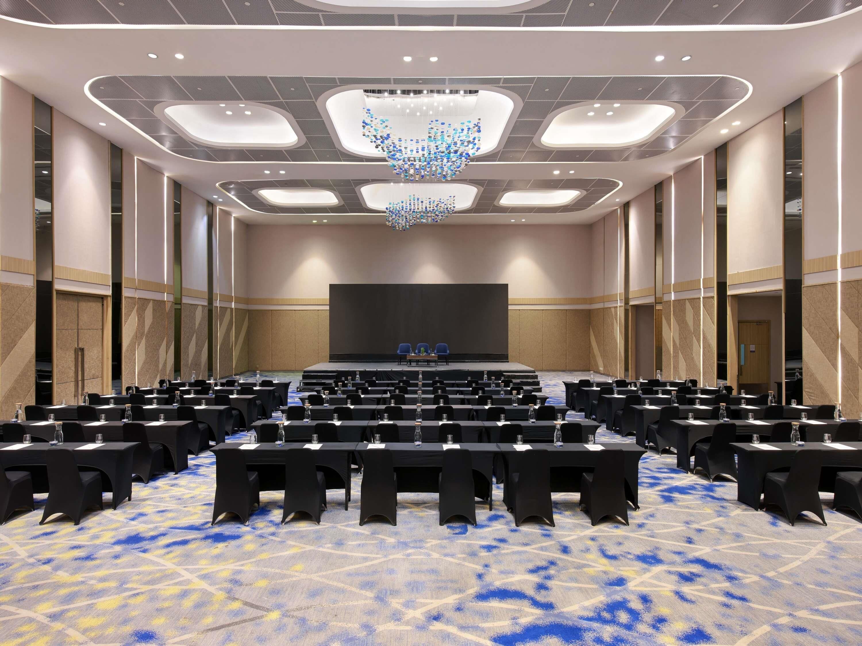A conference room with rows of tables and chairs, a stage, and a large chandelier.