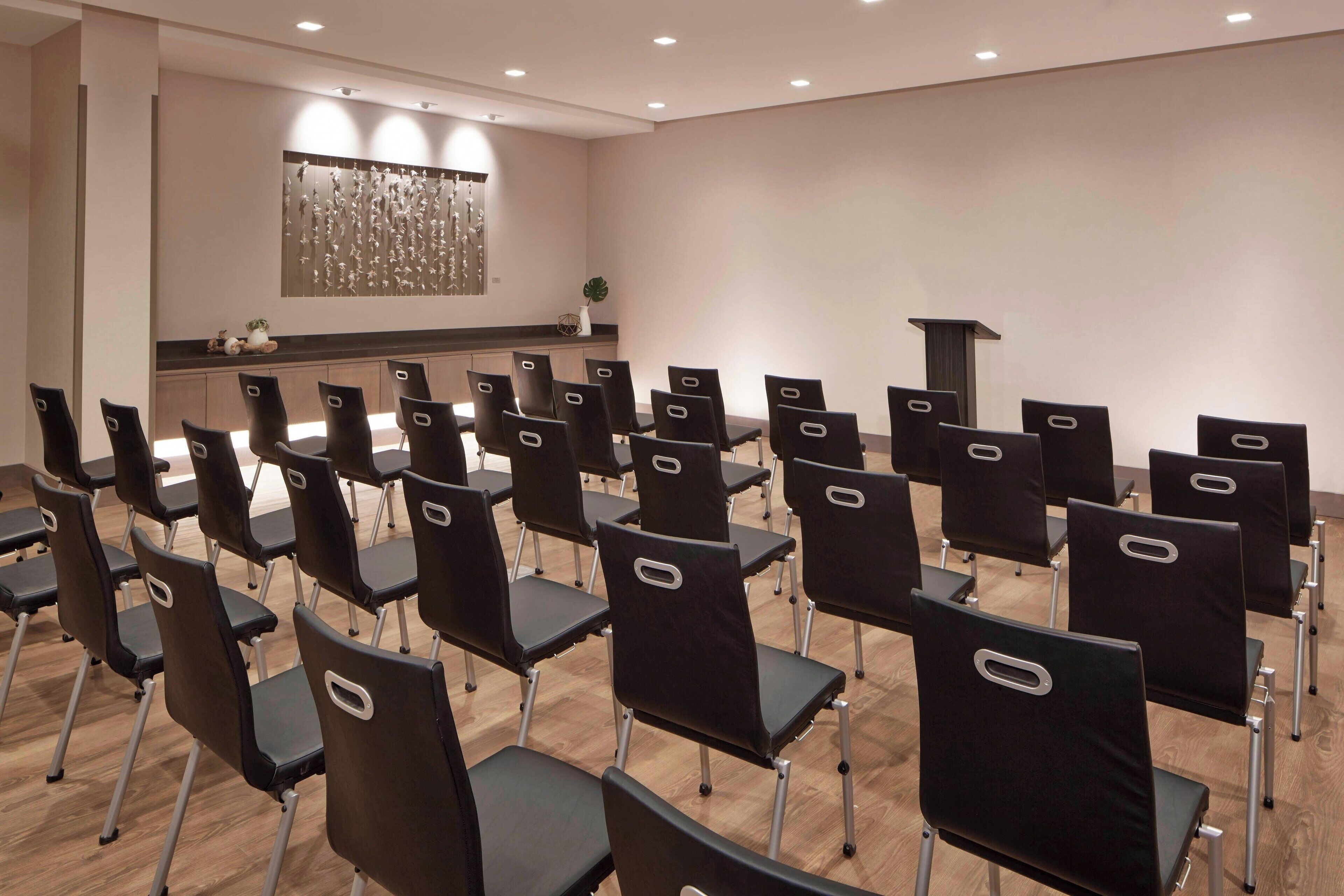 A conference room with rows of black chairs, a wooden floor, a white wall with a decorative panel, and a small plant on a counter.