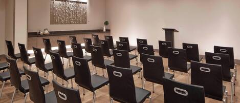 A conference room with rows of black chairs, a wooden floor, a white wall with a decorative panel, and a small plant on a counter.