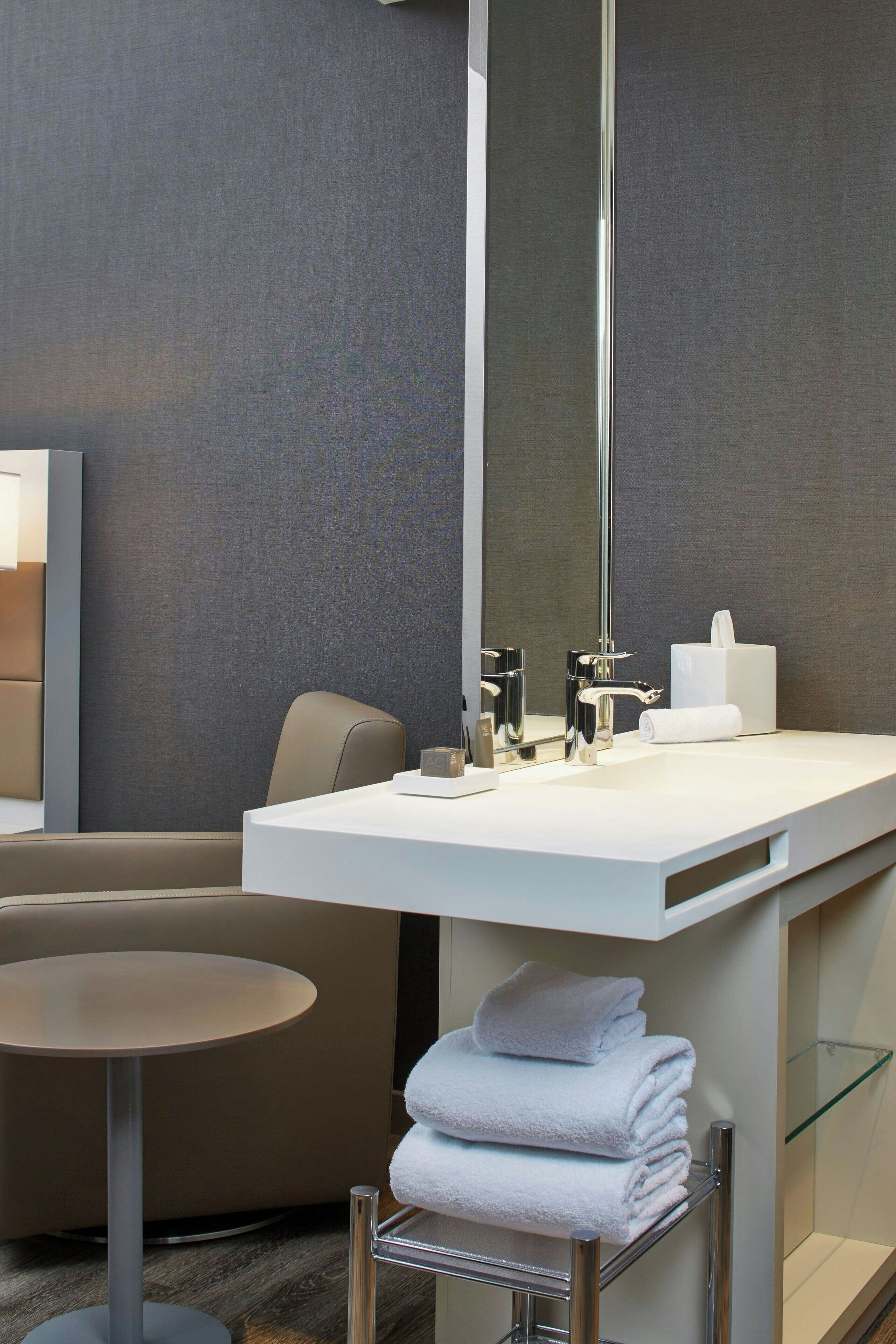 A modern bathroom with a white sink, a large mirror, a towel rack with folded towels, and a round table.