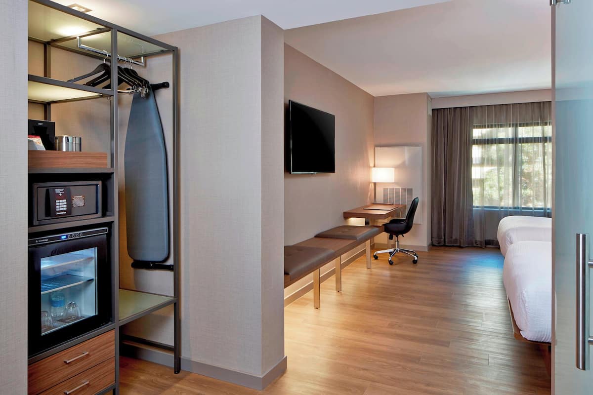 A hotel room with a bed, a desk with a chair, a TV, a mini-fridge, and a wardrobe with a mirror.