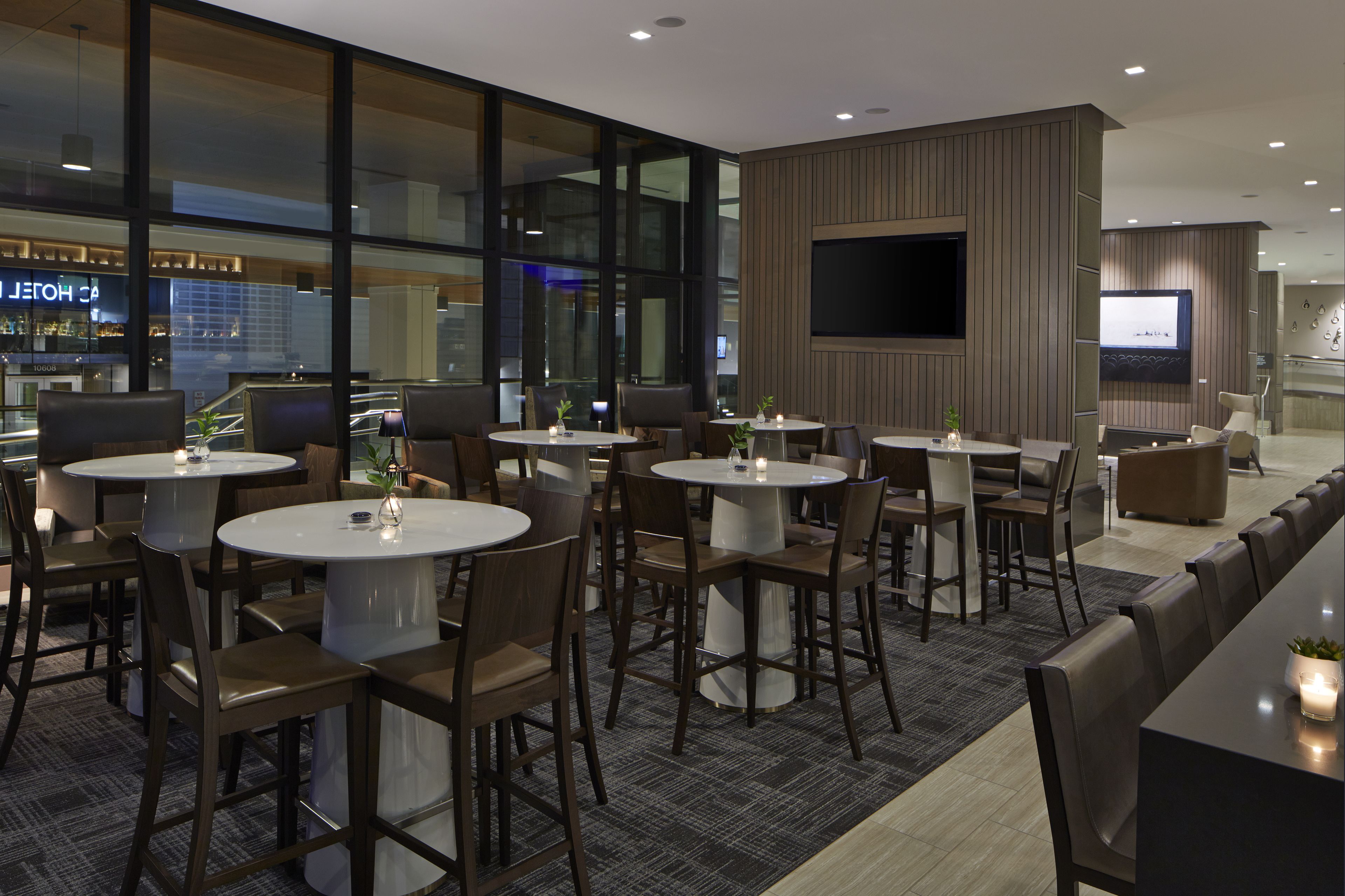 Un vestíbul d'hotel modern amb taules i cadires rodones, una pantalla de televisió gran i vistes a una zona de bar.