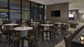 Un vestíbul d'hotel modern amb taules i cadires rodones, una pantalla de televisió gran i vistes a una zona de bar.