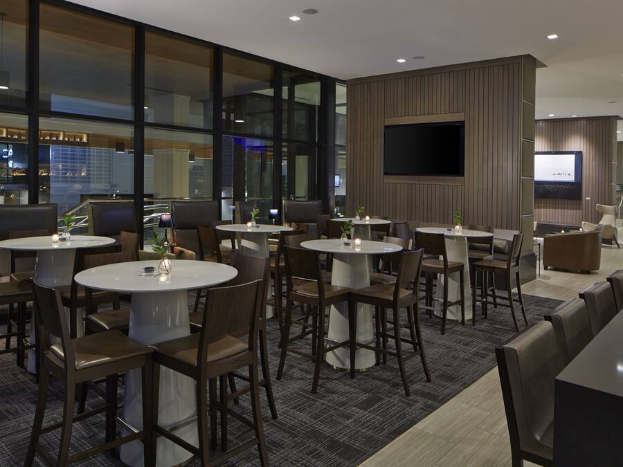 Un vestíbul d'hotel modern amb taules i cadires rodones, una pantalla de televisió gran i vistes a una zona de bar.