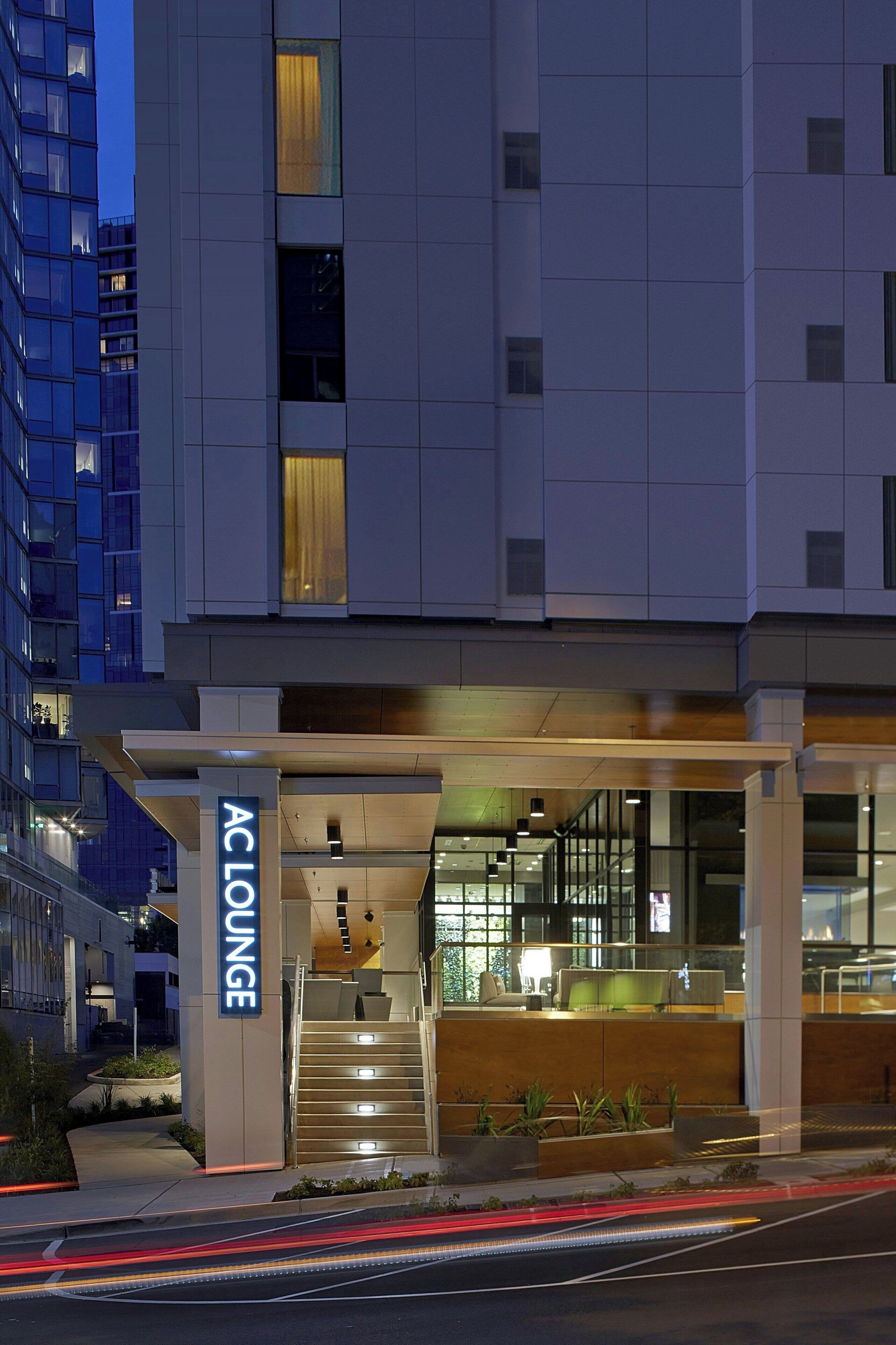 A modern hotel entrance with a sign reading "AC LOUNGE" and a staircase leading to the lobby.
