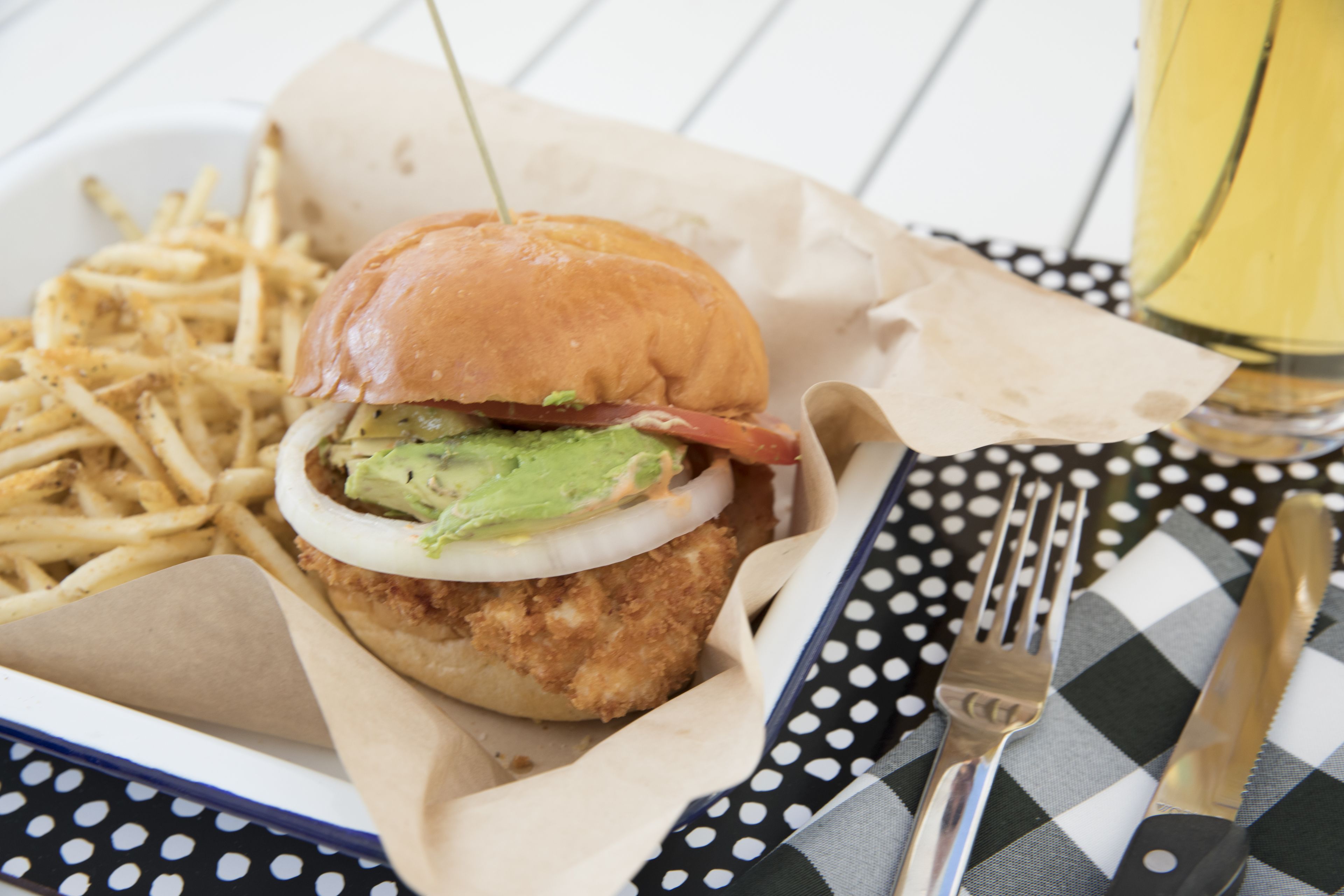 A meal consisting of a breaded chicken sandwich with avocado and onion, accompanied by French fries, on a checkered tablecloth with a fork and knife.