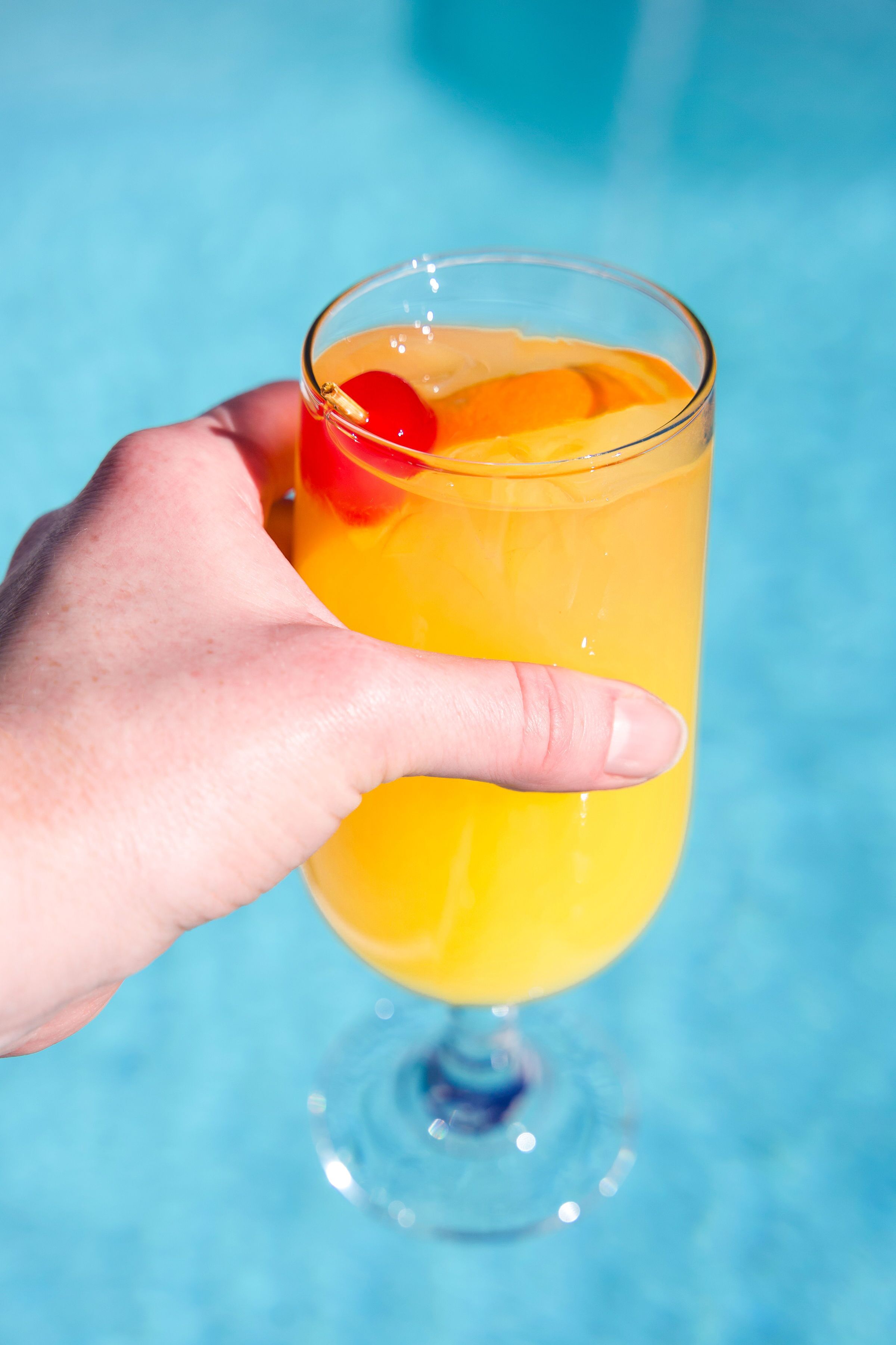 A hand holding a glass of orange drink with a cherry garnish, against a blue background.
