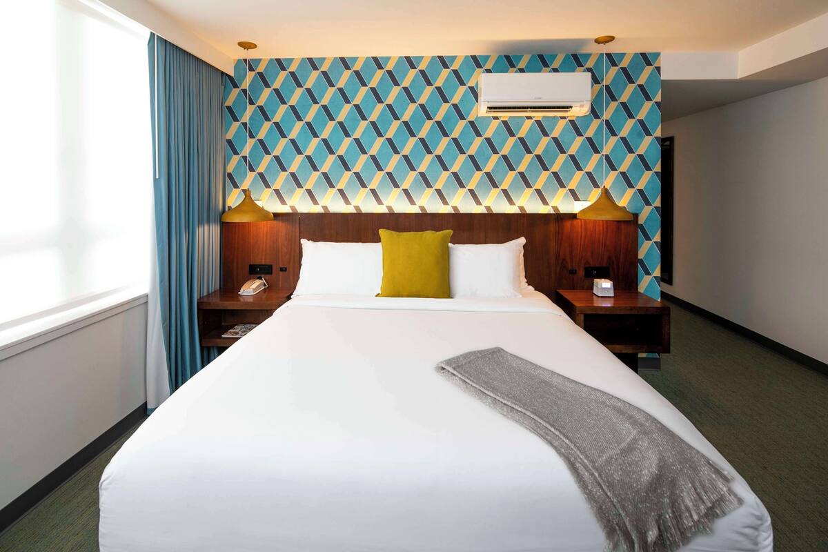 A hotel room with a large bed, a wooden headboard, two bedside tables with lamps, a wall-mounted air conditioner, and a patterned wallpaper.
