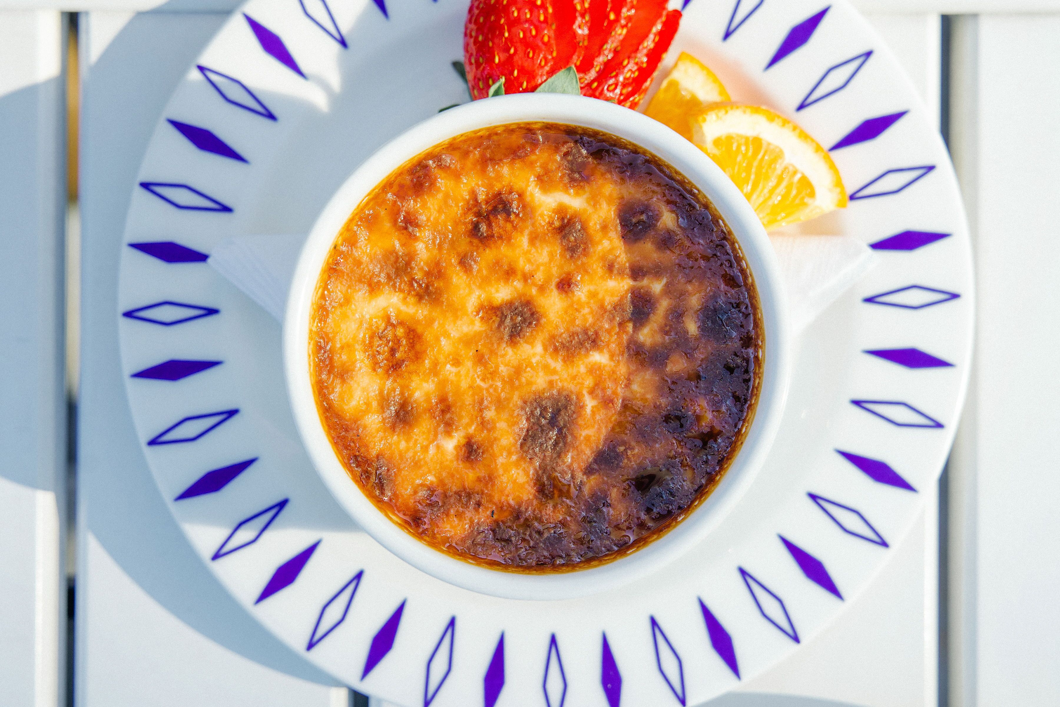 A bowl of crème brûlée on a white plate with a blue pattern, accompanied by a strawberry and an orange slice.