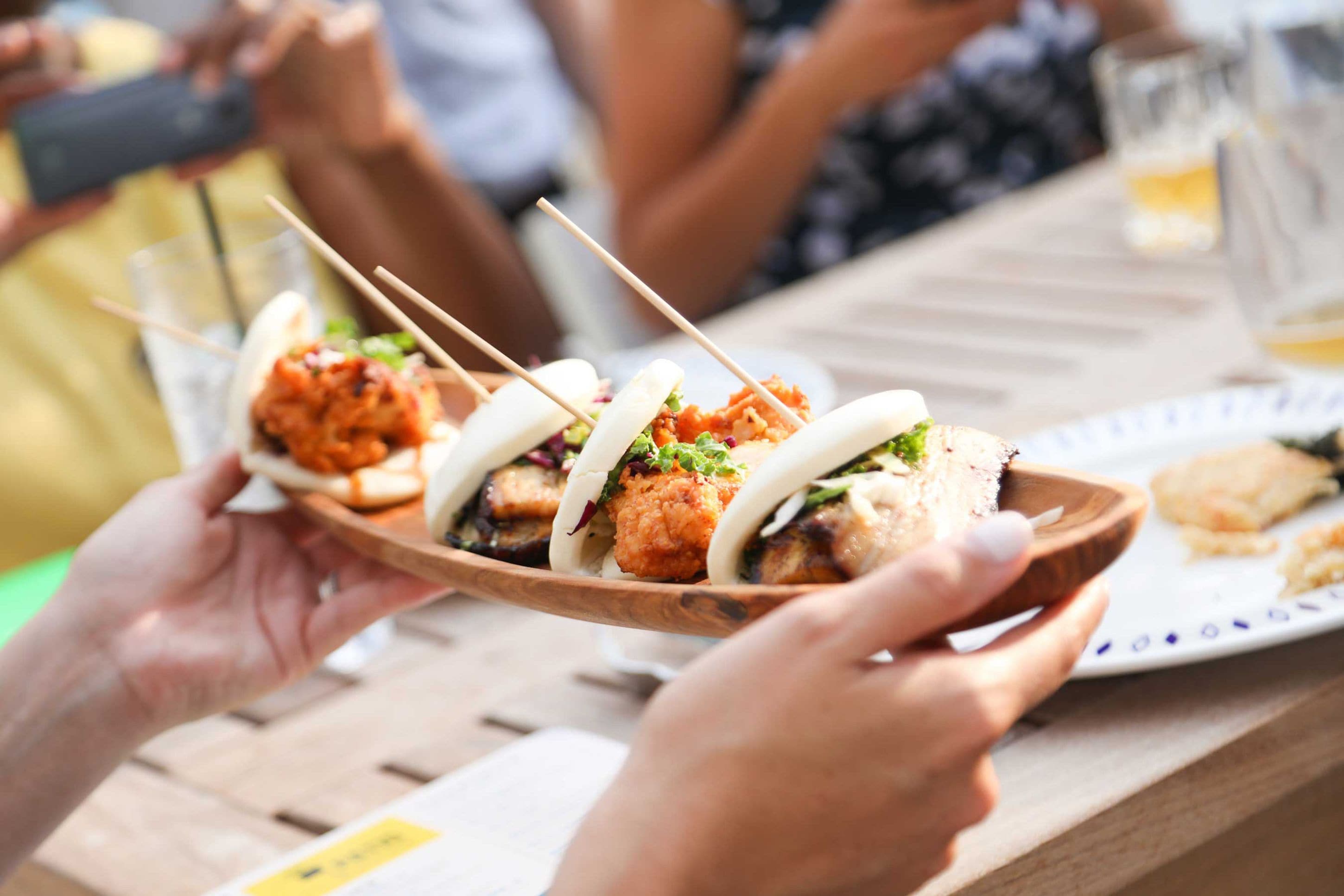 A wooden serving tray with skewered food items, held by hands at a table.