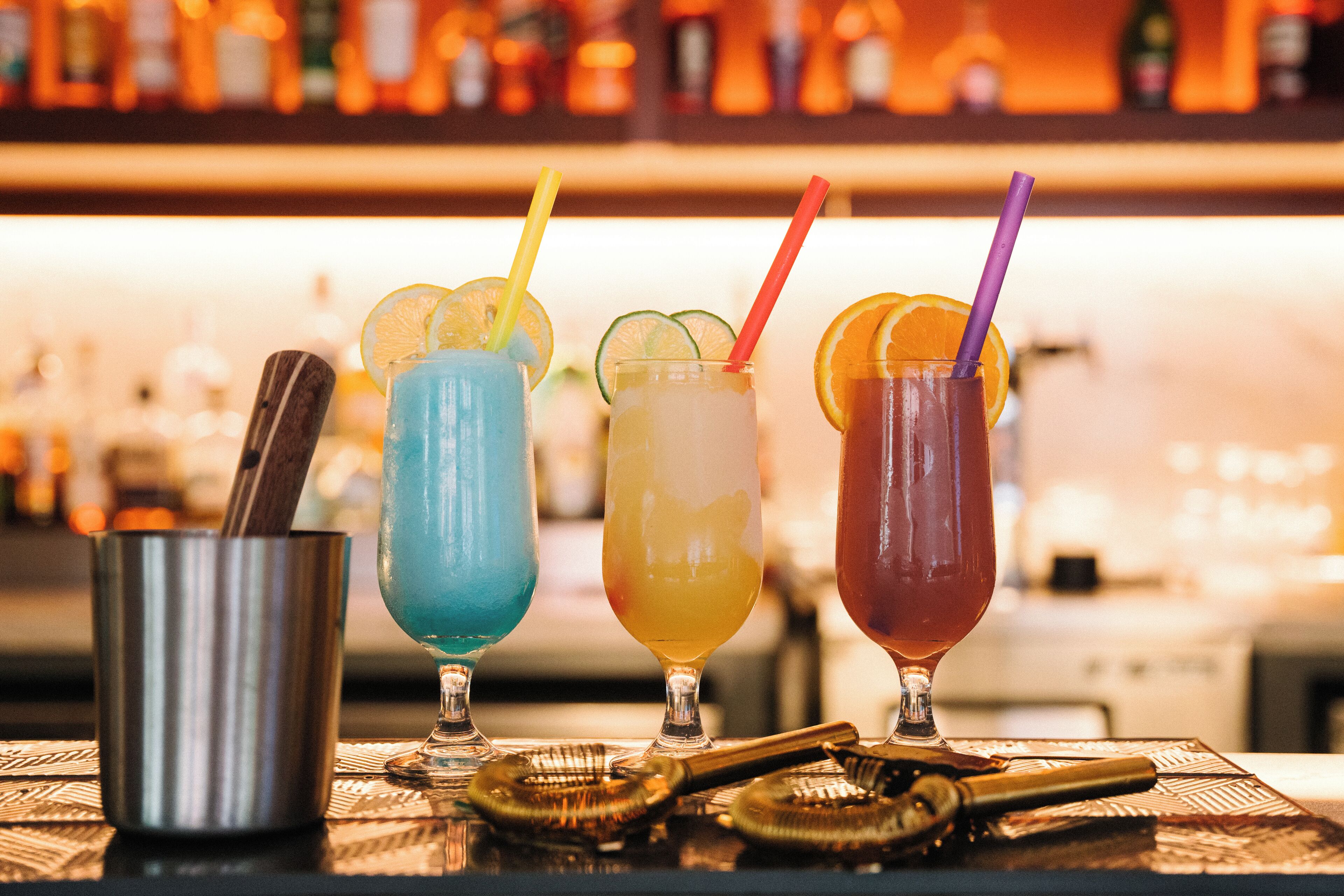 A bar setting with four colorful cocktails, a metal cup, and cocktail shakers on a patterned tablecloth.