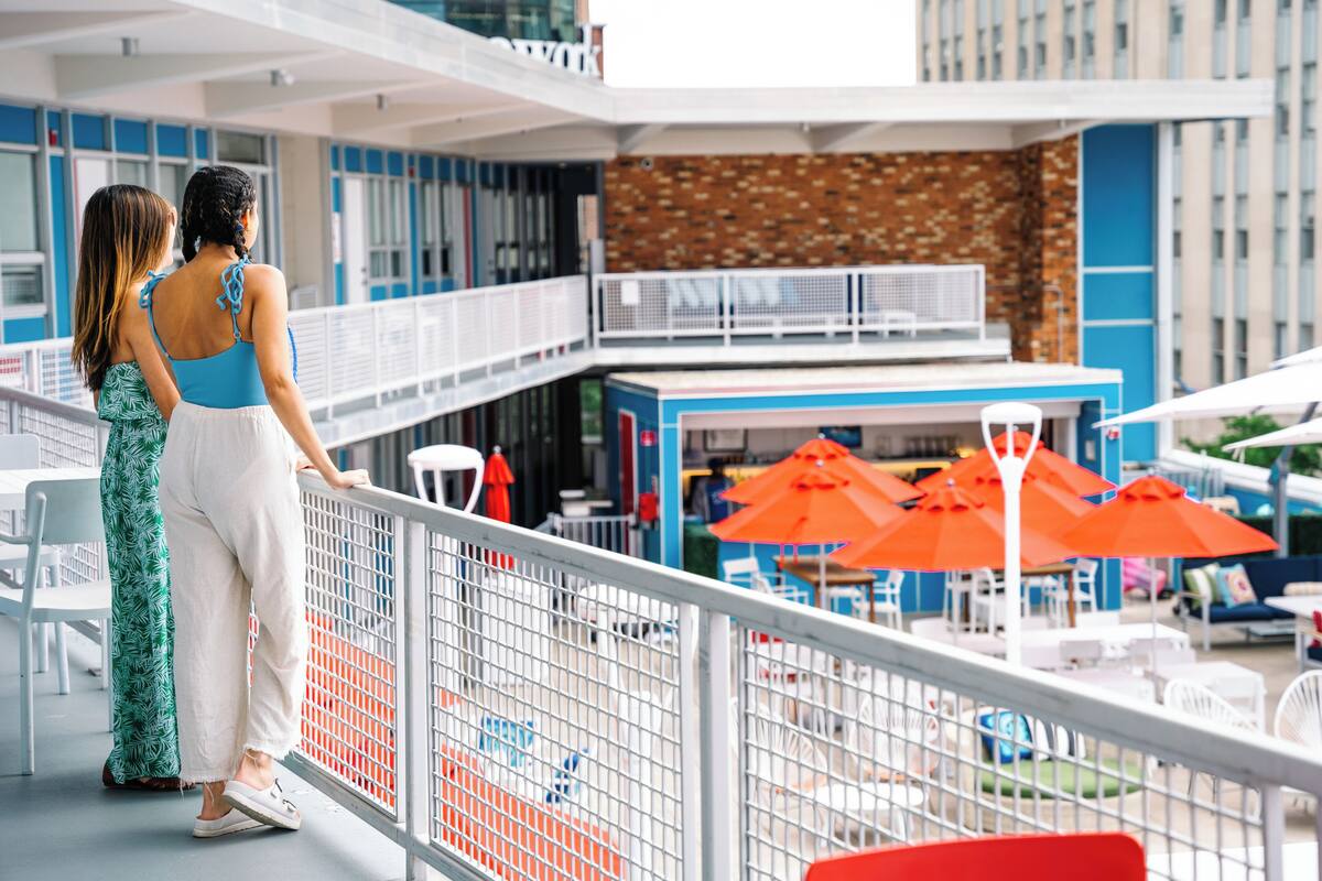 A balcony with two people overlooking a dining area with orange umbrellas.