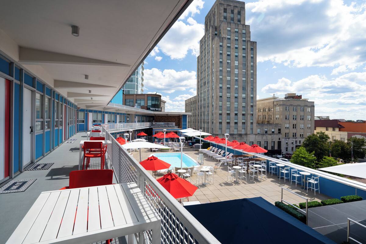 A rooftop pool area with red umbrellas, white lounge chairs, and a view of a tall building in the background.