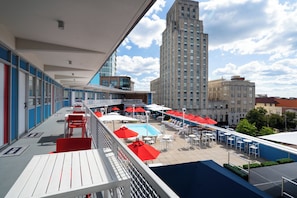 A rooftop pool area with red umbrellas, white lounge chairs, and a view of a tall building in the background.