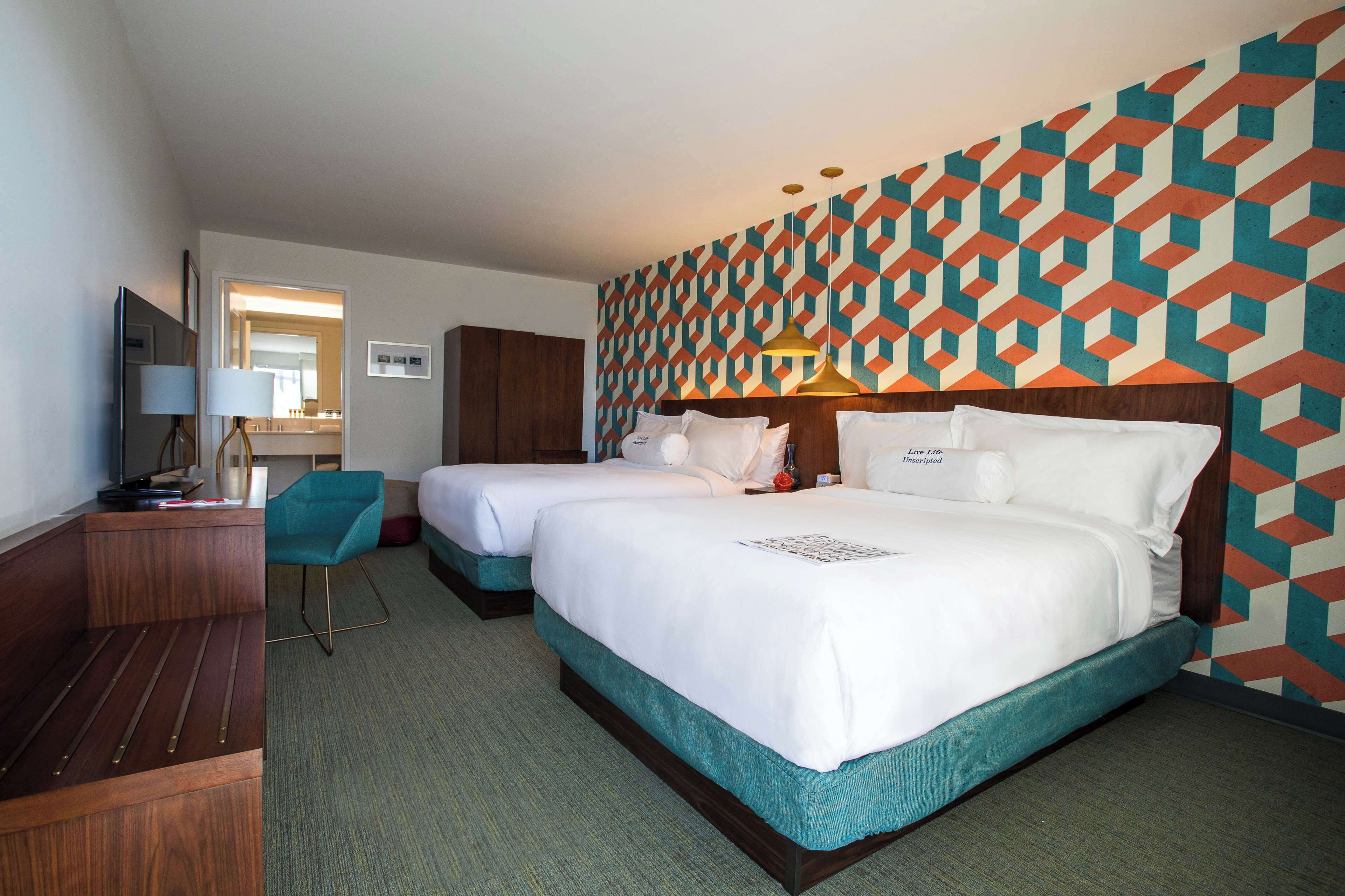 A hotel room with two beds, a desk, a chair, and a patterned wall.