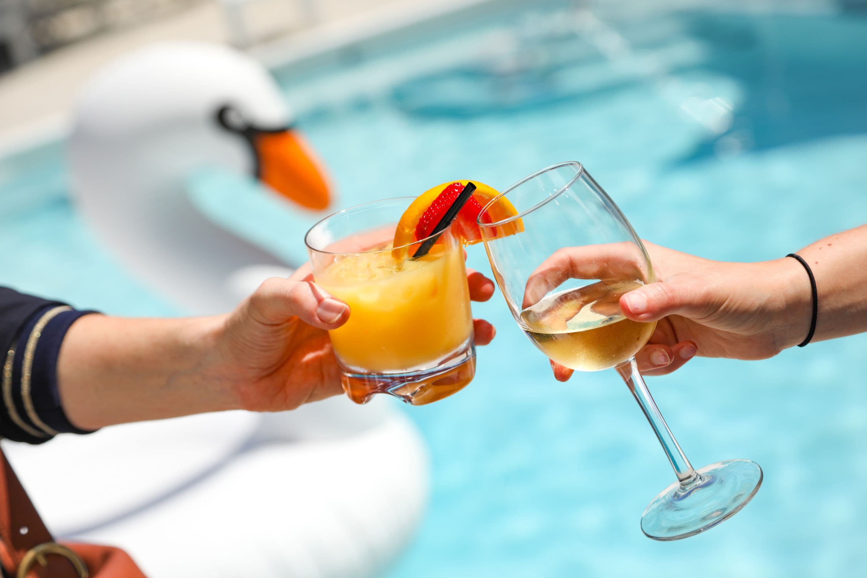 Two people holding glasses of drinks by a pool with a swan float.
