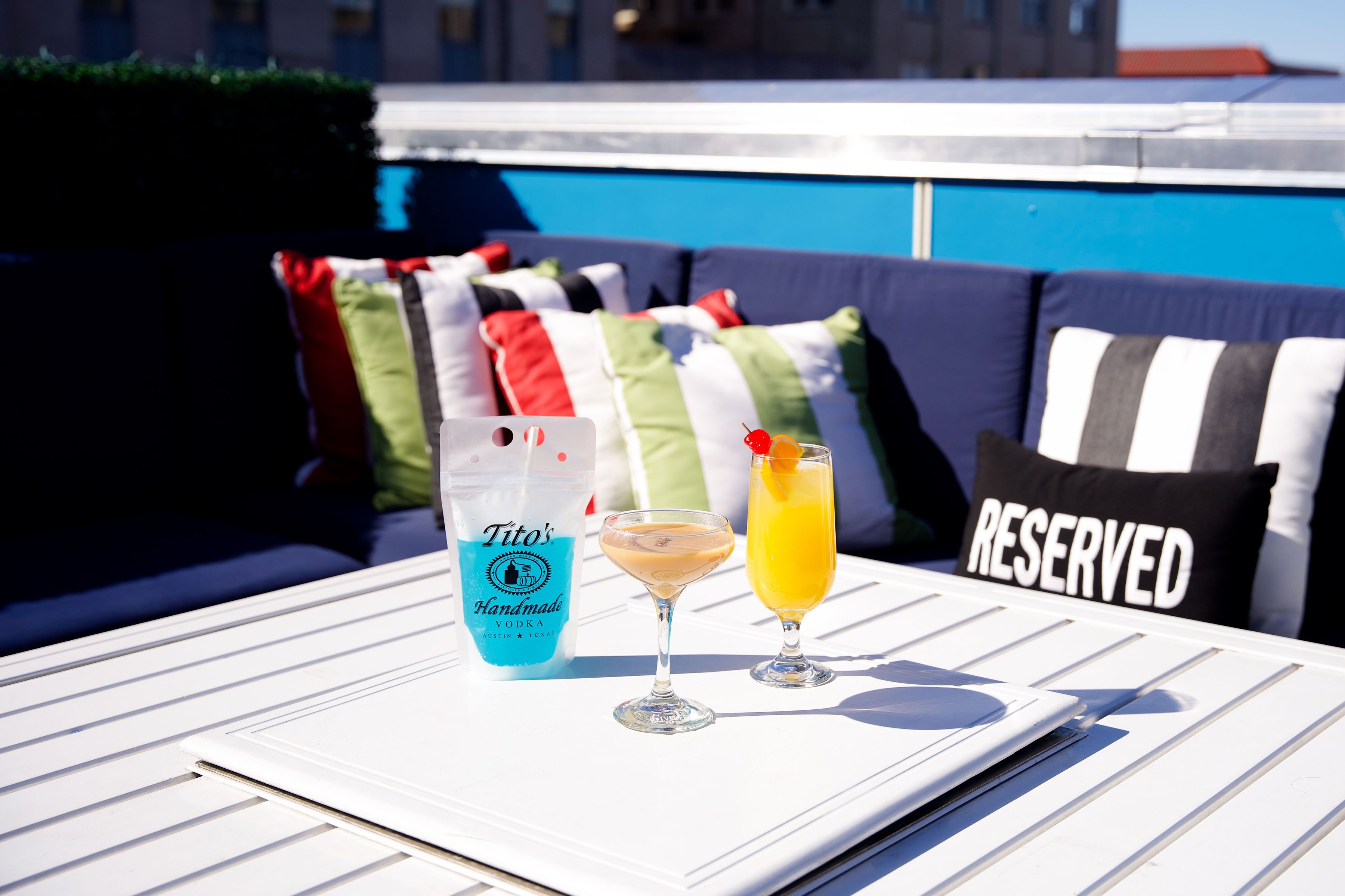 A white table with a bottle of Tito's Handmade Vodka and two drinks, a martini and a cocktail, on a rooftop terrace with a pool and colorful cushions in the background.