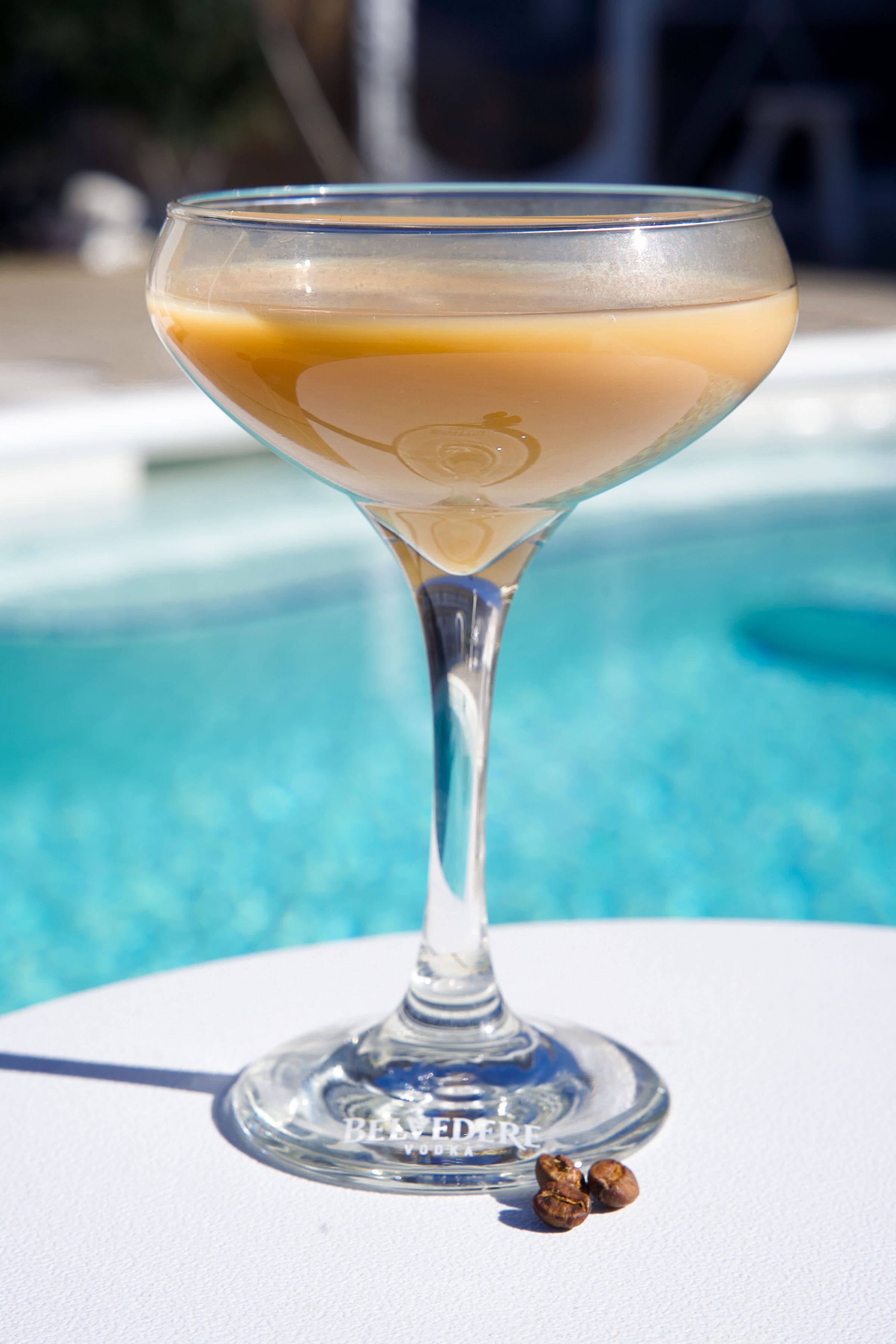 A clear glass cocktail with a stem, placed on a white surface near a swimming pool.