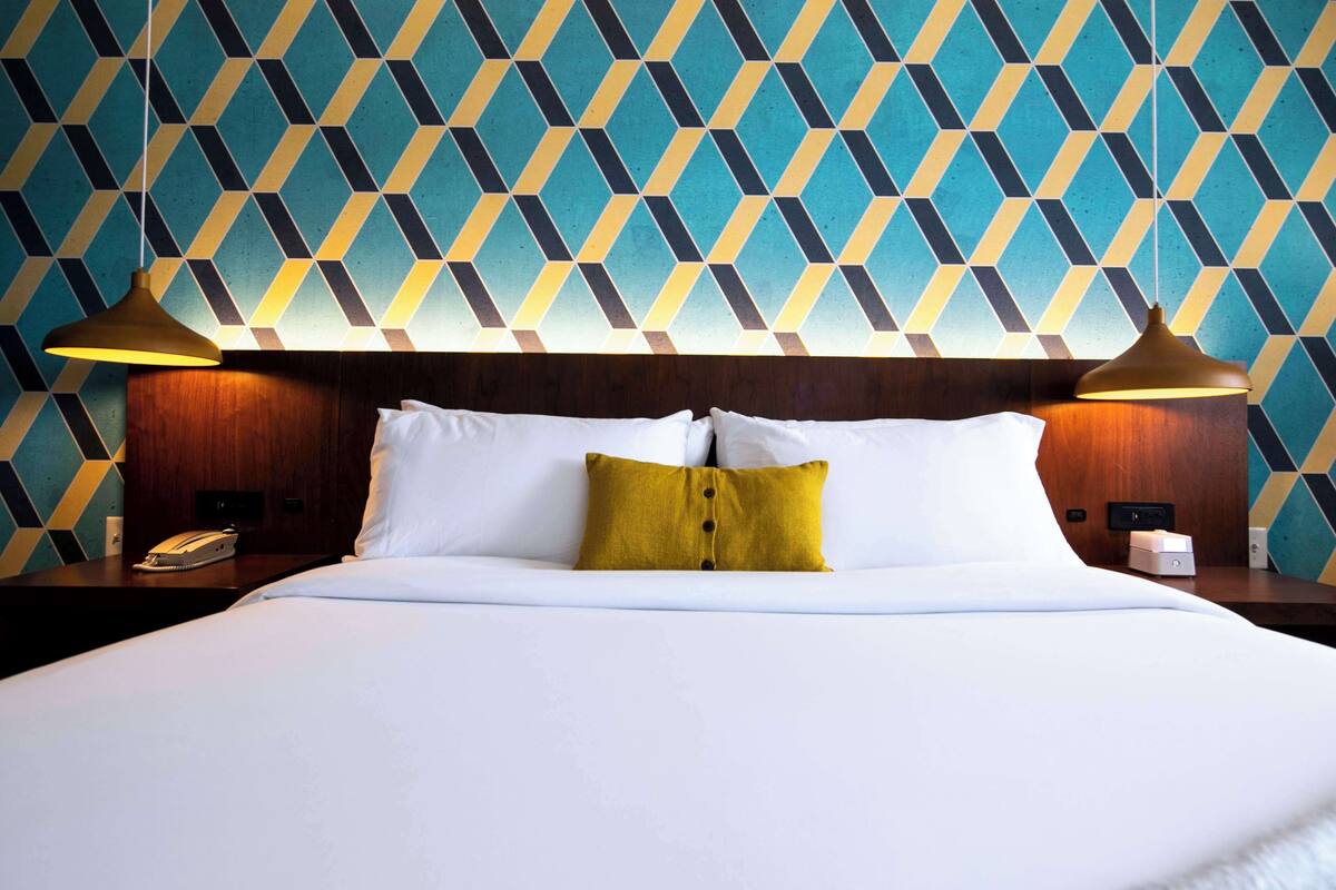 A hotel room with a patterned wall, a bed with white and yellow pillows, and two bedside lamps.