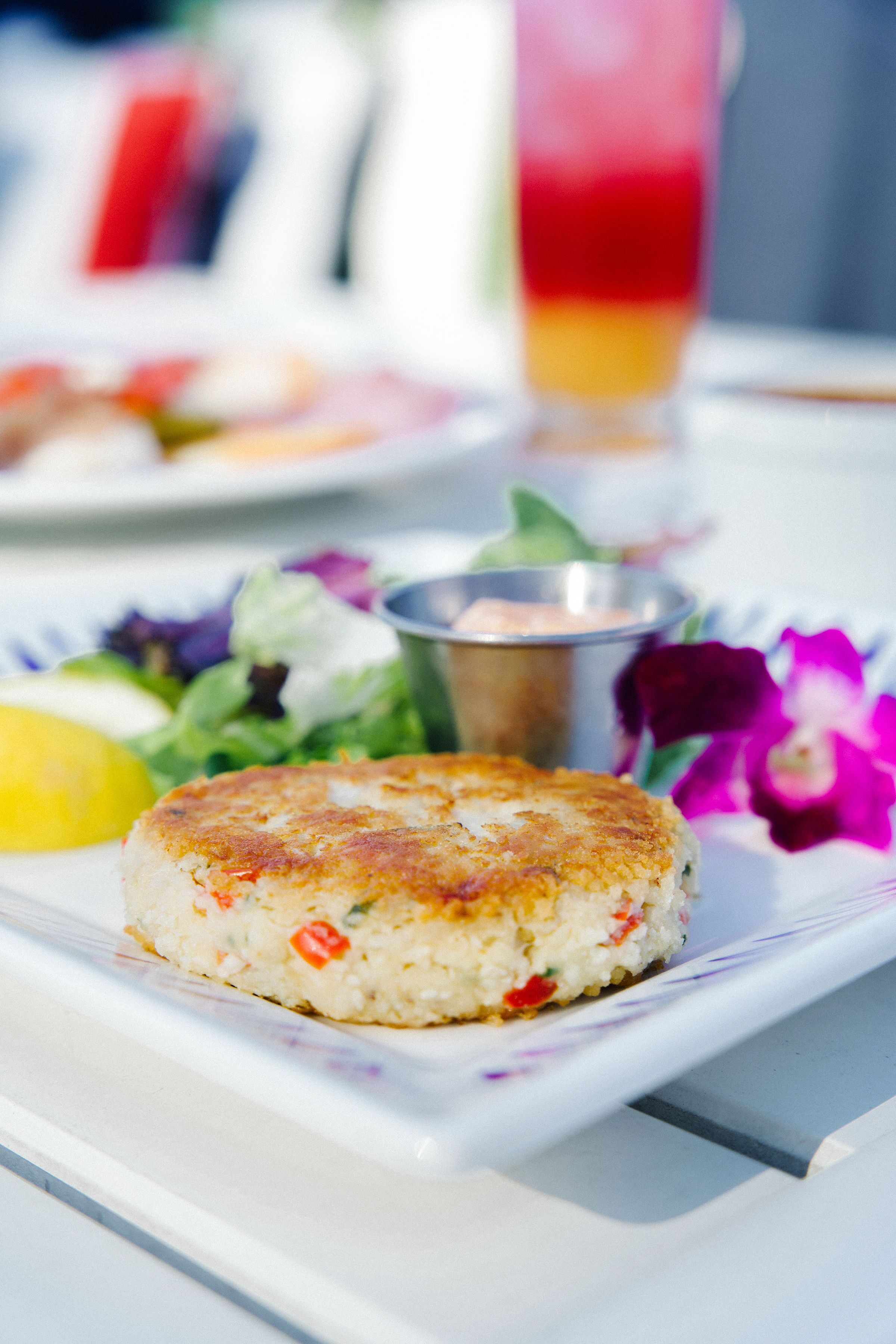 A plate with a crab cake and a side salad, a glass of drink, and a small metal container on a table.