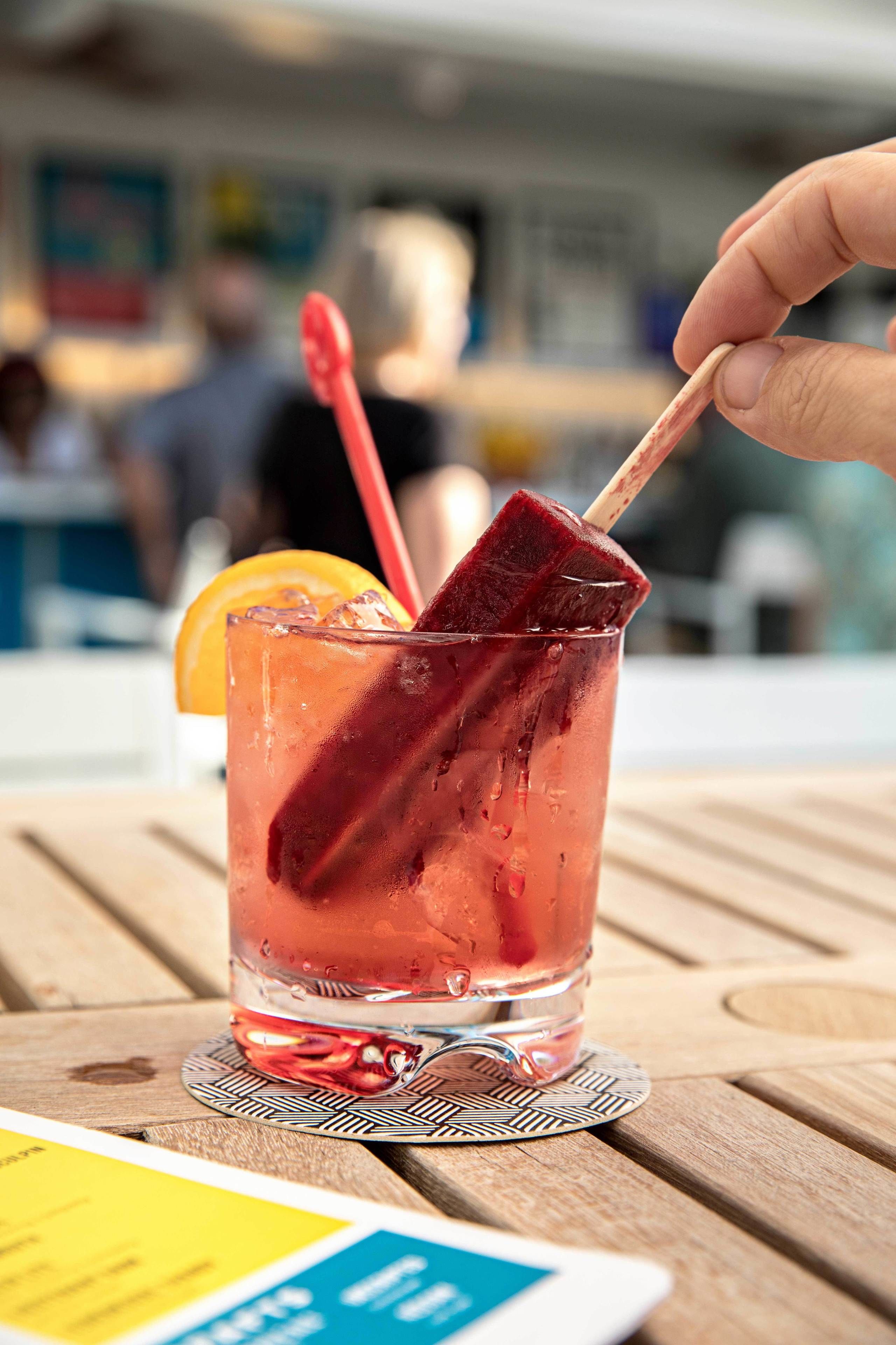 A glass of iced drink with a red straw and a slice of fruit on a wooden table.