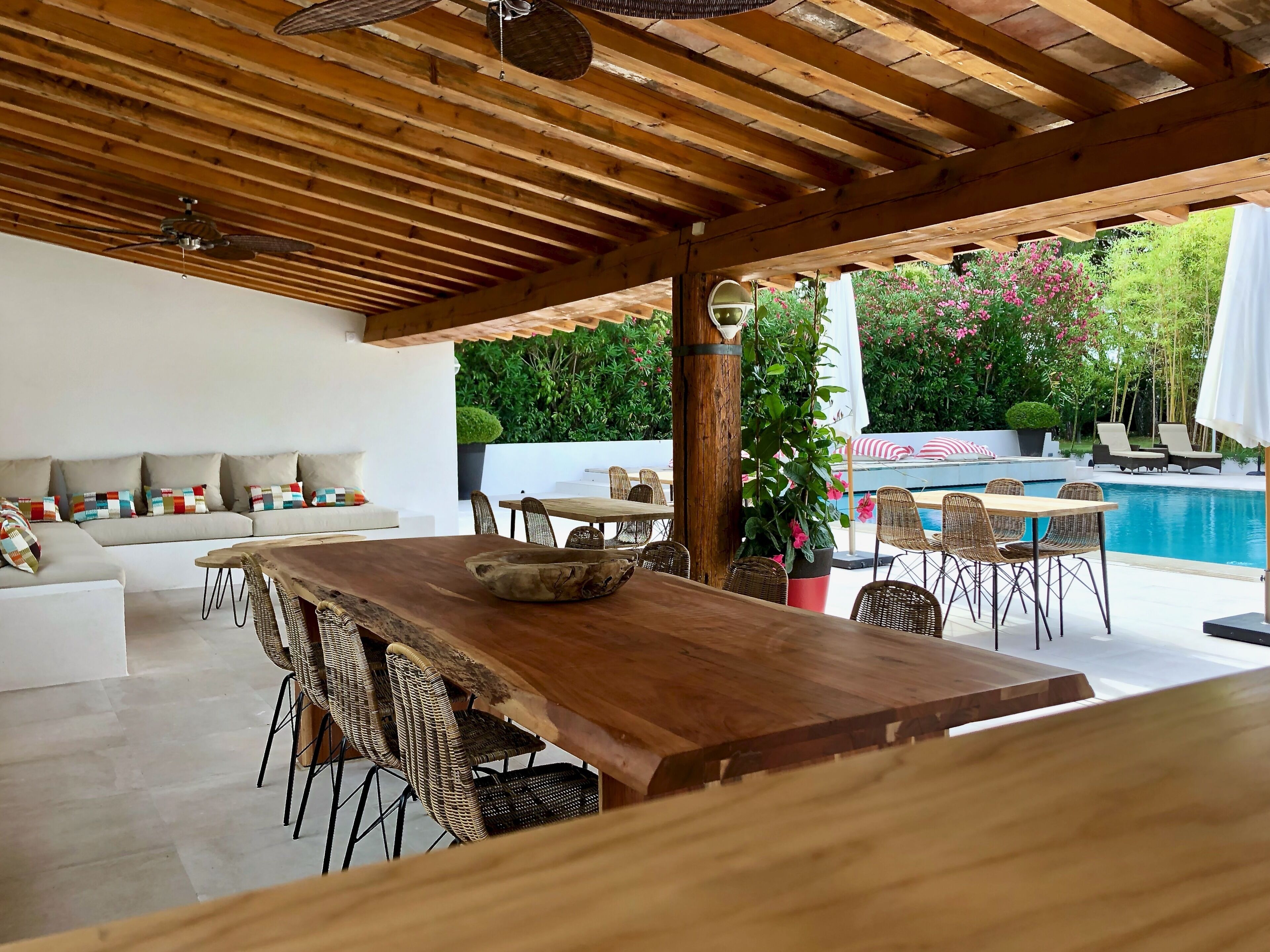 Un patio al aire libre con una mesa y sillas de madera, una piscina y una zona de estar con cojines.