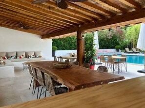 An outdoor patio with a wooden table and chairs, a pool, and a seating area with cushions.