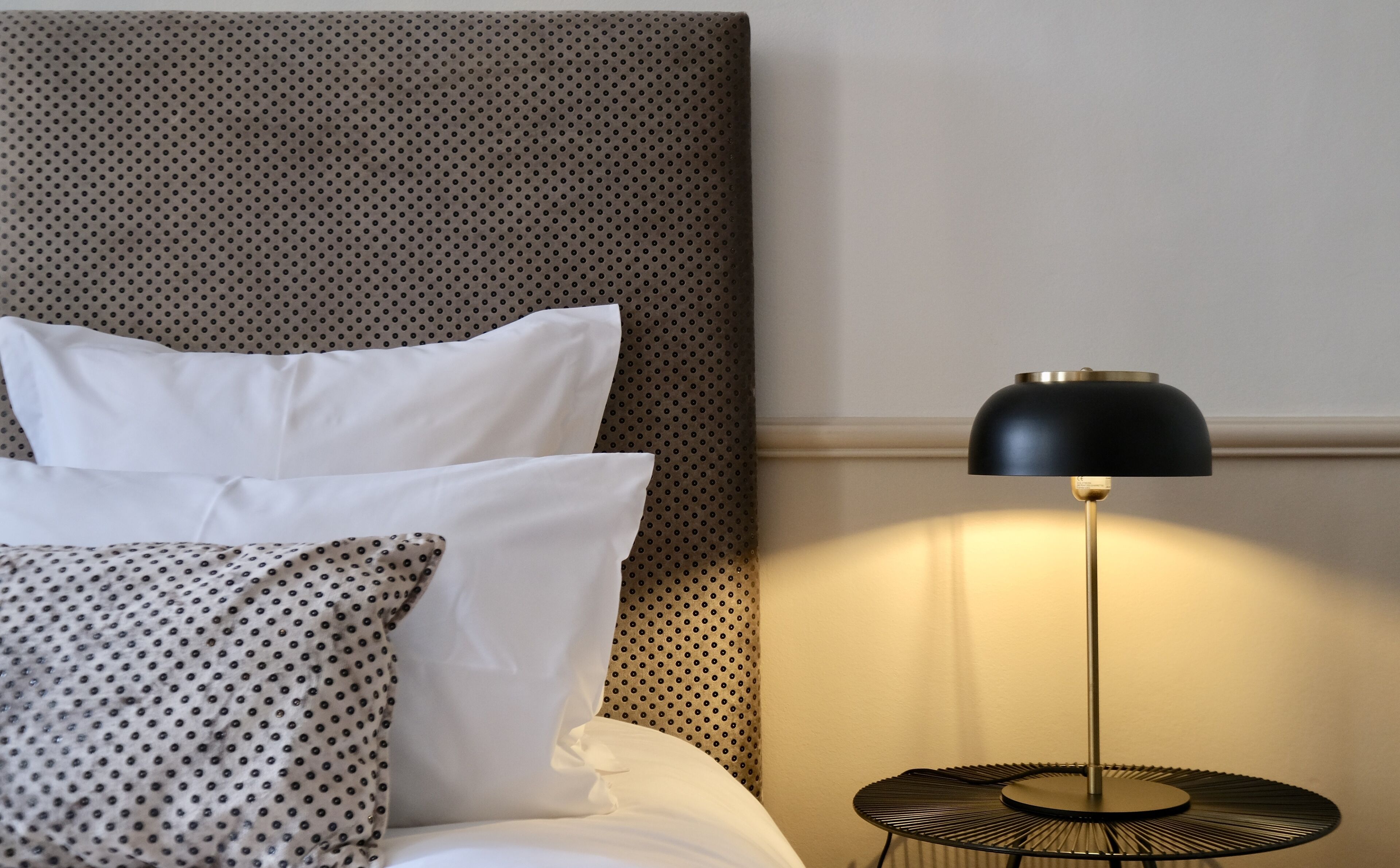 A neatly made bed with white and patterned pillows, a black lamp on a small table beside it, and a textured wall behind.