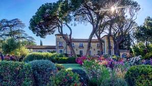 Un edificio grande e histórico con fachada simétrica, rodeado de un jardín bien cuidado con arbustos y plantas de flores diversas.